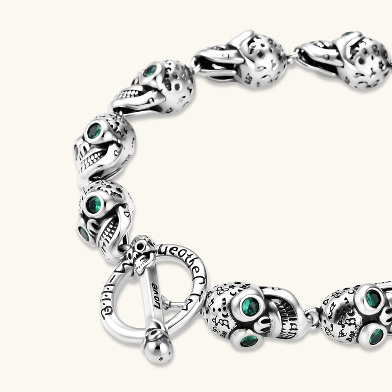 Silver stainless steel skull link bracelet with green crystal eye sockets and engraved detailing, toggle clasp closure.