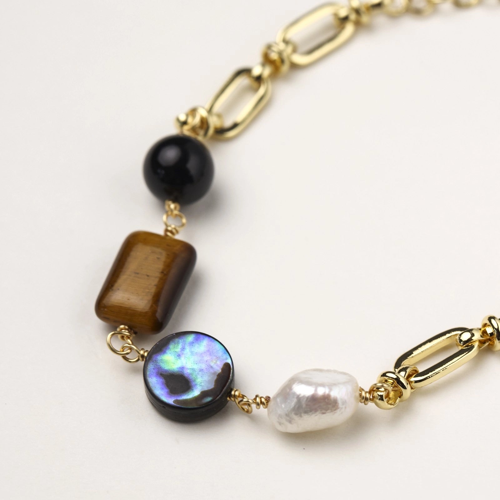 Gold-plated chain bracelet featuring tiger's eye, obsidian, freshwater pearls and abalone shell on white background