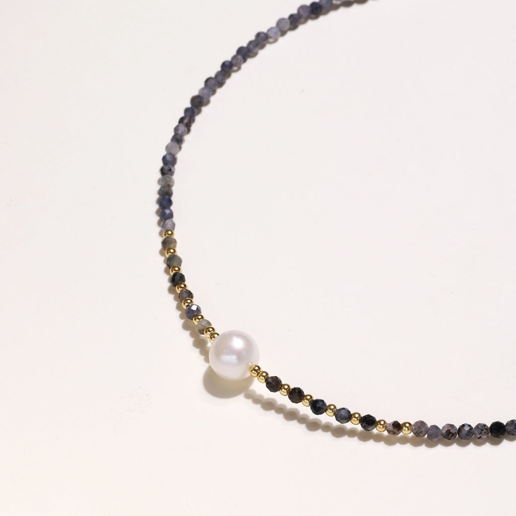 Delicate beaded necklace with violet iolite stones, gold spacers, and central white freshwater pearl on neutral background.