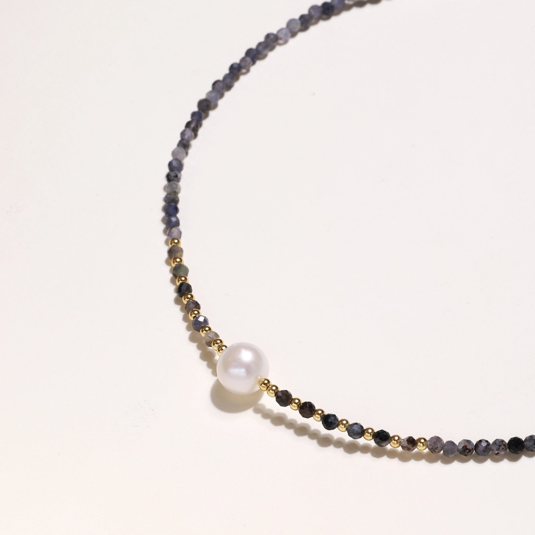 Delicate beaded necklace with violet iolite stones, gold spacers, and central white freshwater pearl on neutral background.