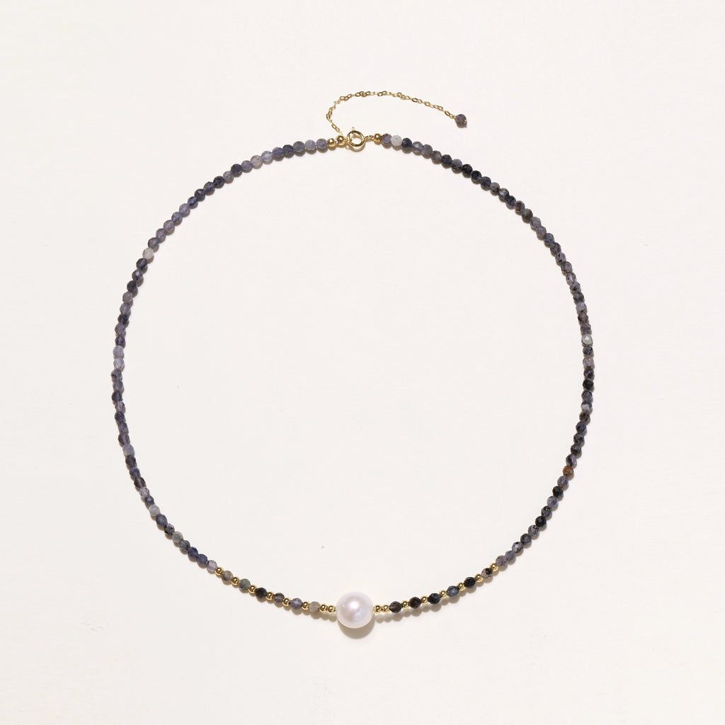 Delicate beaded necklace with violet iolite stones, gold spacers, and central white freshwater pearl on neutral background.